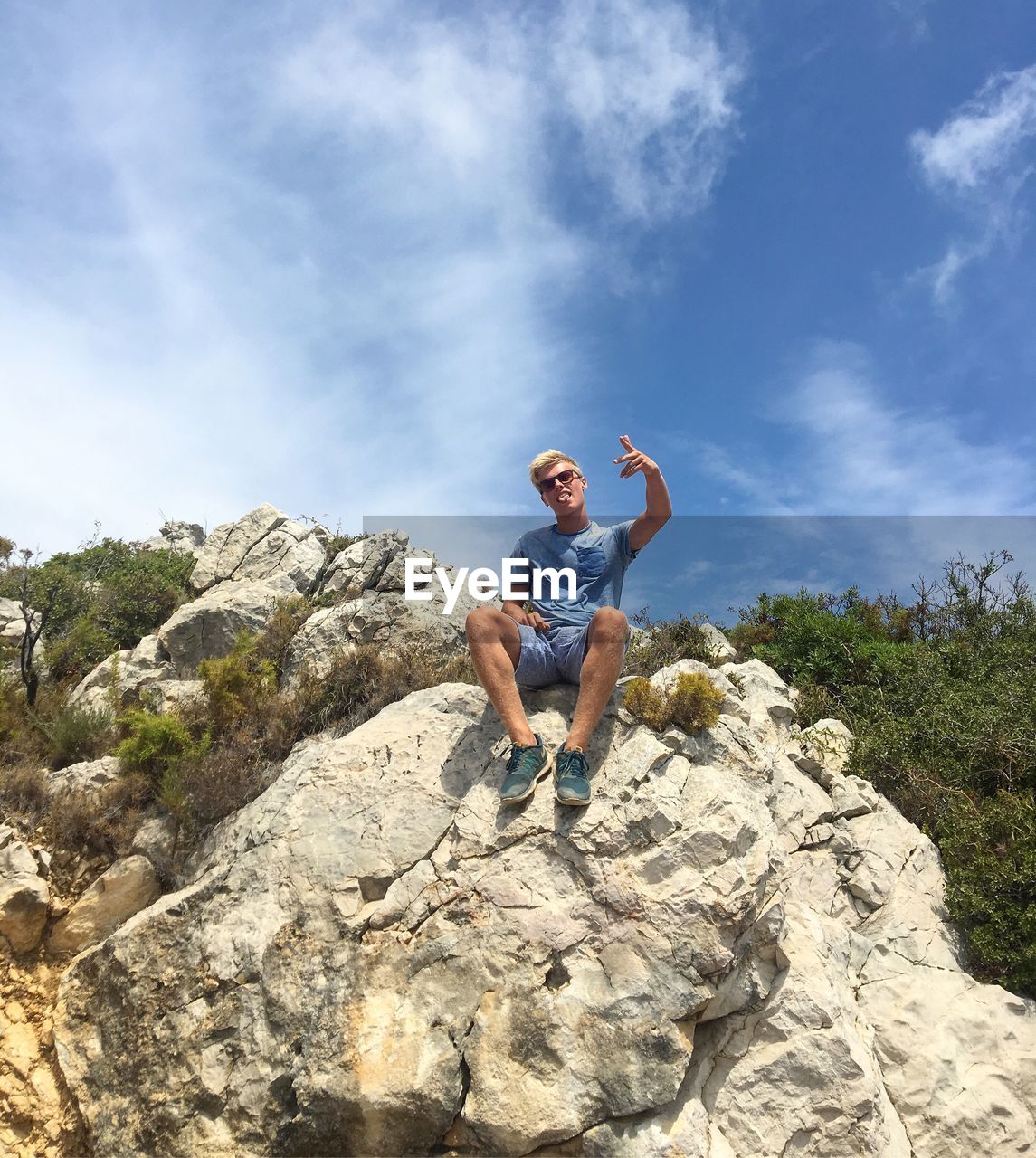 Portrait of man gesturing while sitting on rocks against sky