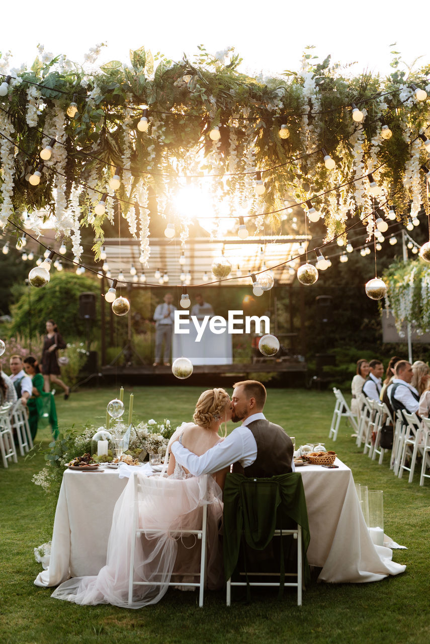 wedding, ceremony, event, wedding dress, celebration, plant, adult, togetherness, life events, women, love, men, nature, bride, table, food and drink, positive emotion, wedding reception, emotion, married, group of people, party, enjoyment, social gathering, newlywed, sitting, crowd, chair, grass, front or back yard, relaxation, happiness, female, seat, wedding ceremony, lifestyles, young adult, tree, bonding, romance, friendship, leisure activity, summer, backyard, drink, sunlight, full length, outdoors, food, furniture, wedding guest, day, refreshment, copy space, weekend activities, garden party, meal, beginnings, architecture, lawn, glass
