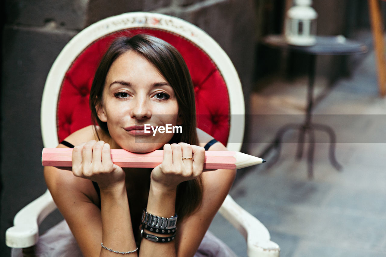 Portrait of beautiful woman holding large pencil while sitting on chair at home