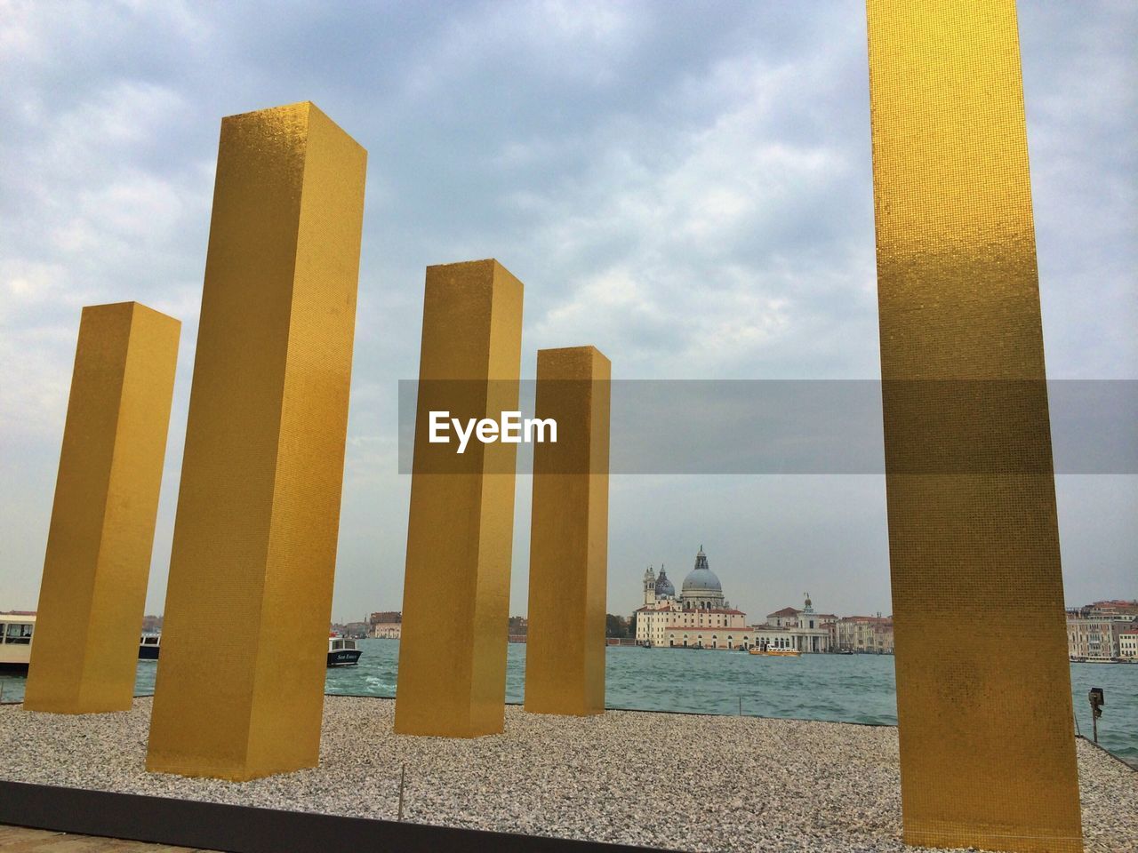 Low angle view of gold colored columns by river and city against cloudy sky