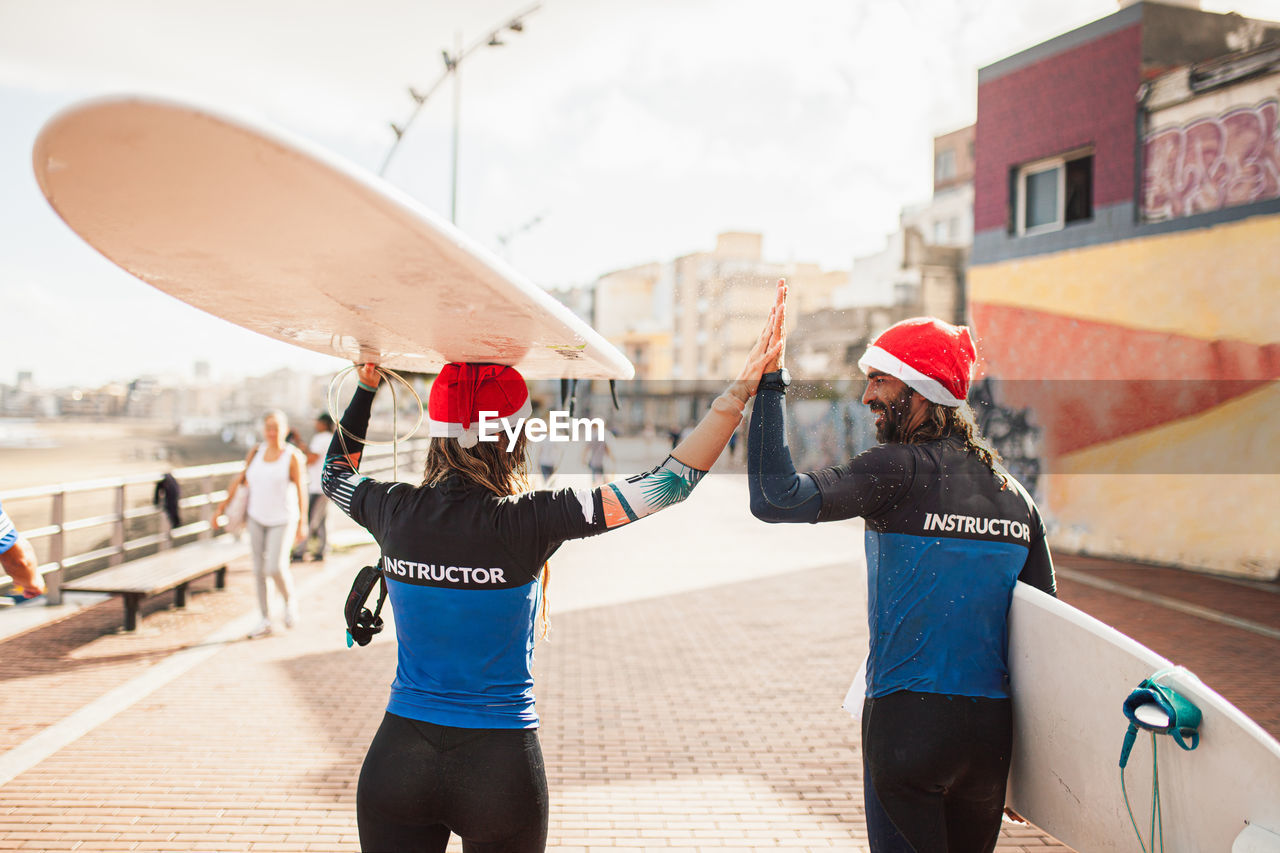 Rear view of couple wearing santa hats with surfboards walking on footpath in city