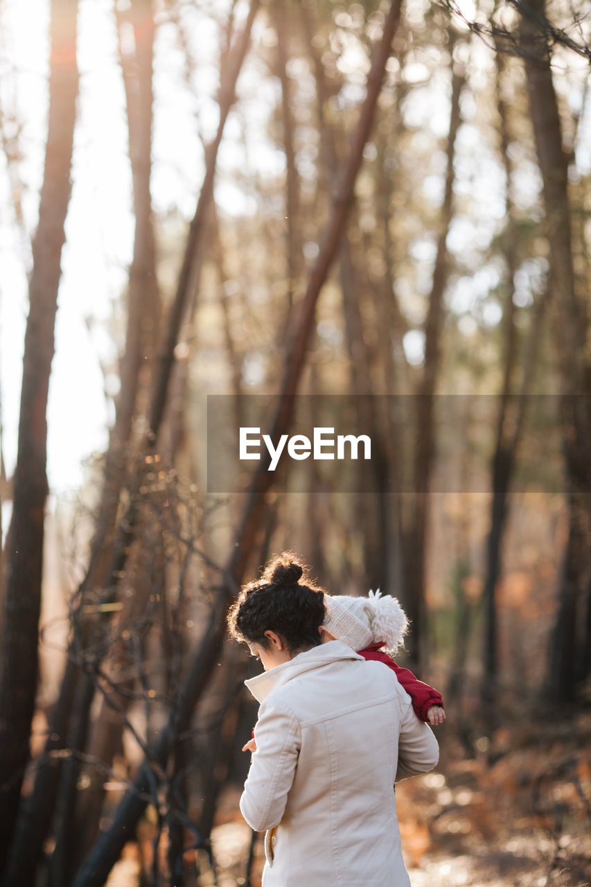 Rear view of mother carrying baby girl standing against trees at forest