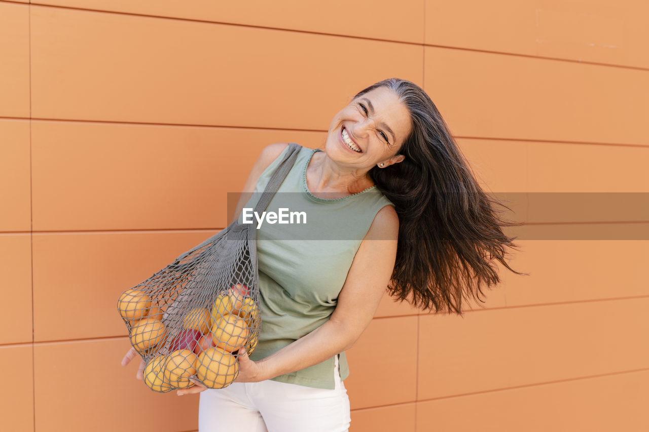 Happy woman with mesh bag of fruits standing by orange wall