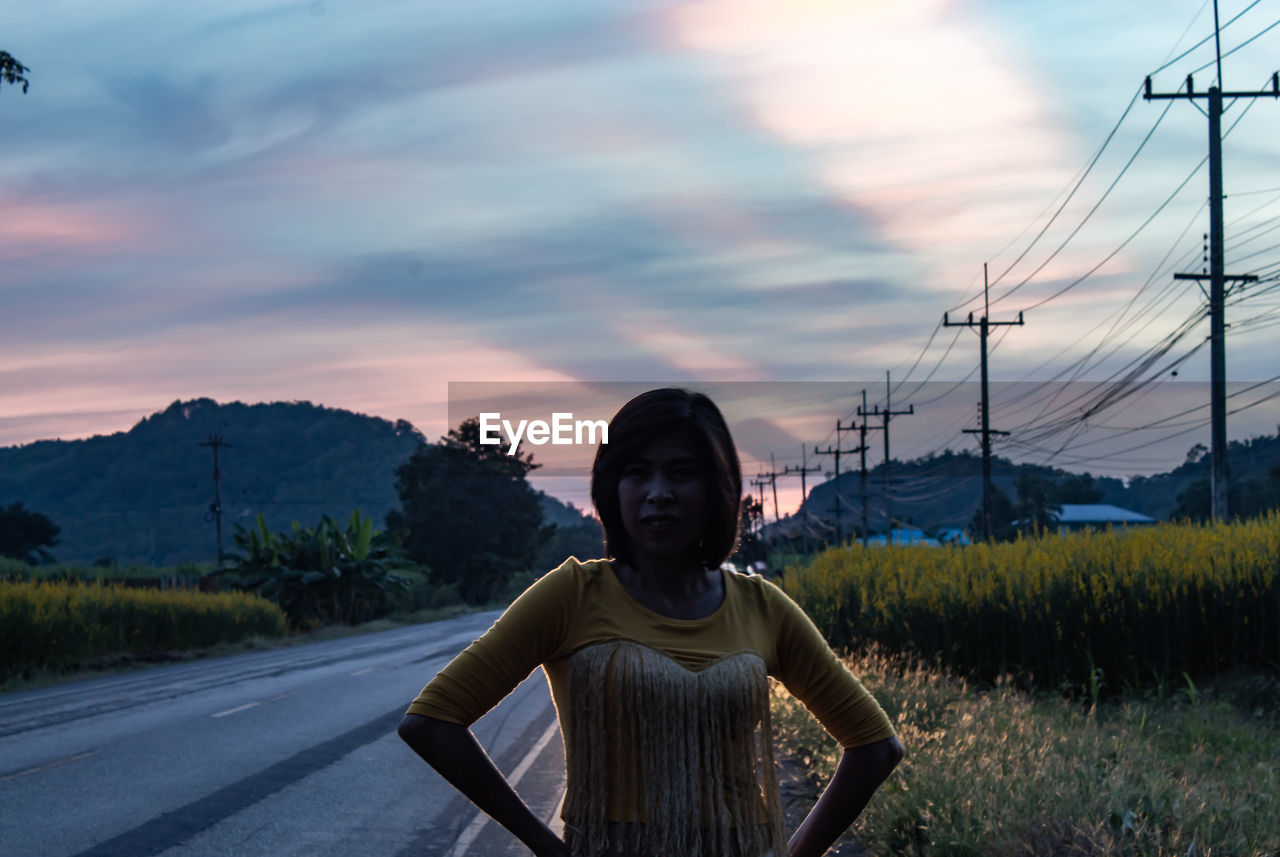 Portrait of woman standing on roadside against sky during sunset