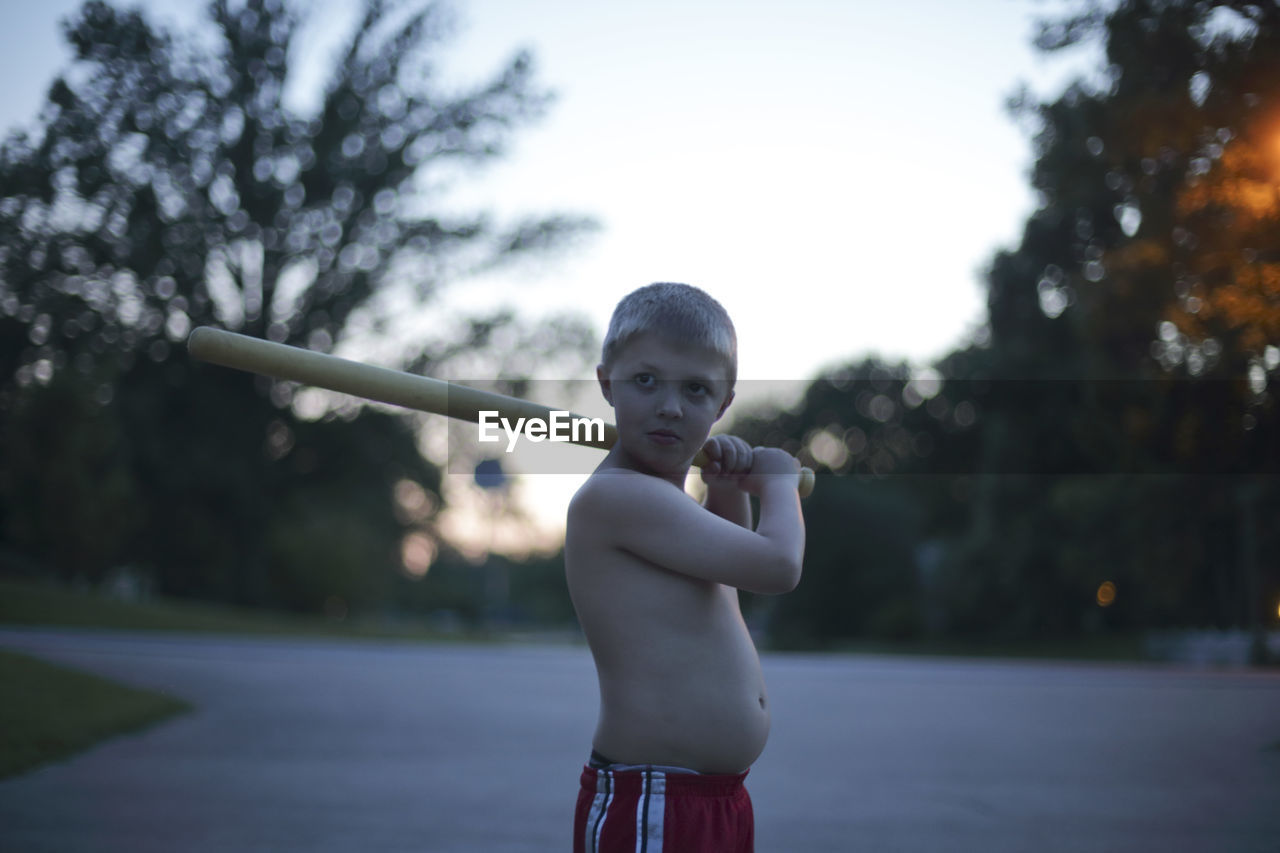 Shirtless boy holding baseball bat against trees at park