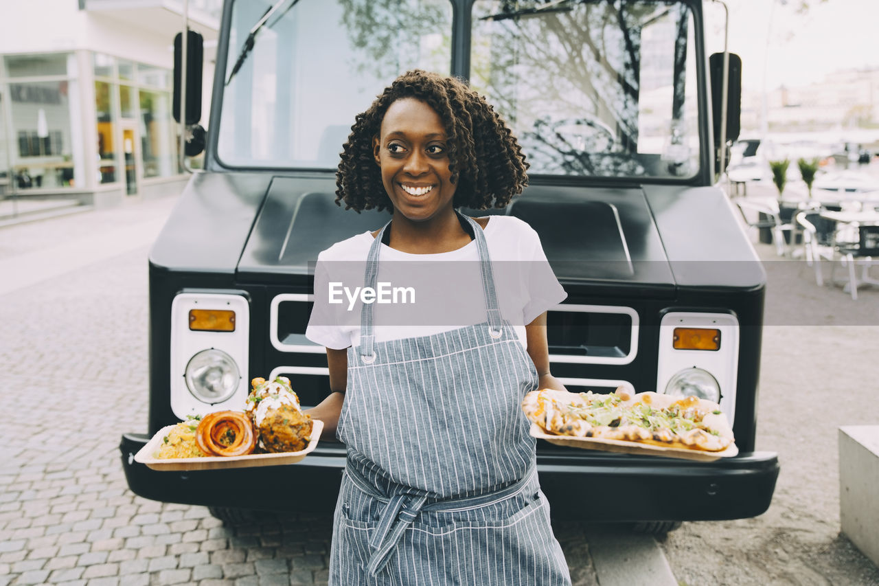 Smiling female owner holding indian food plate while looking away in city