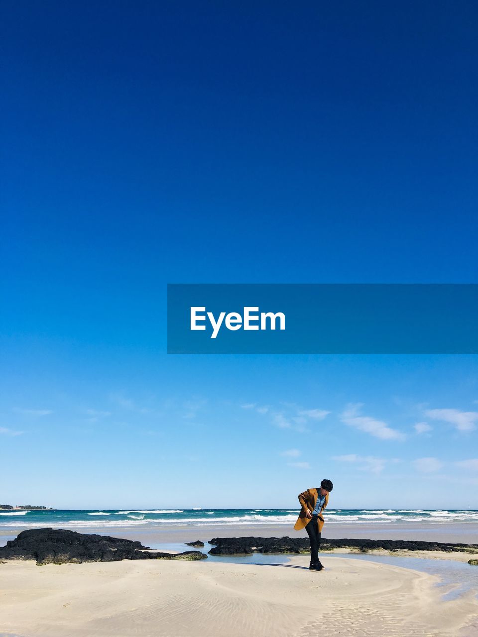 Young man looking down while standing at beach against blue sky