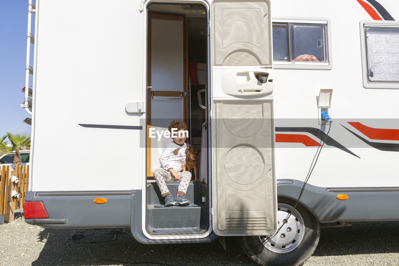 Little curly-haired boy smiling sitting in the door of motor home, tenderly holding his pet.