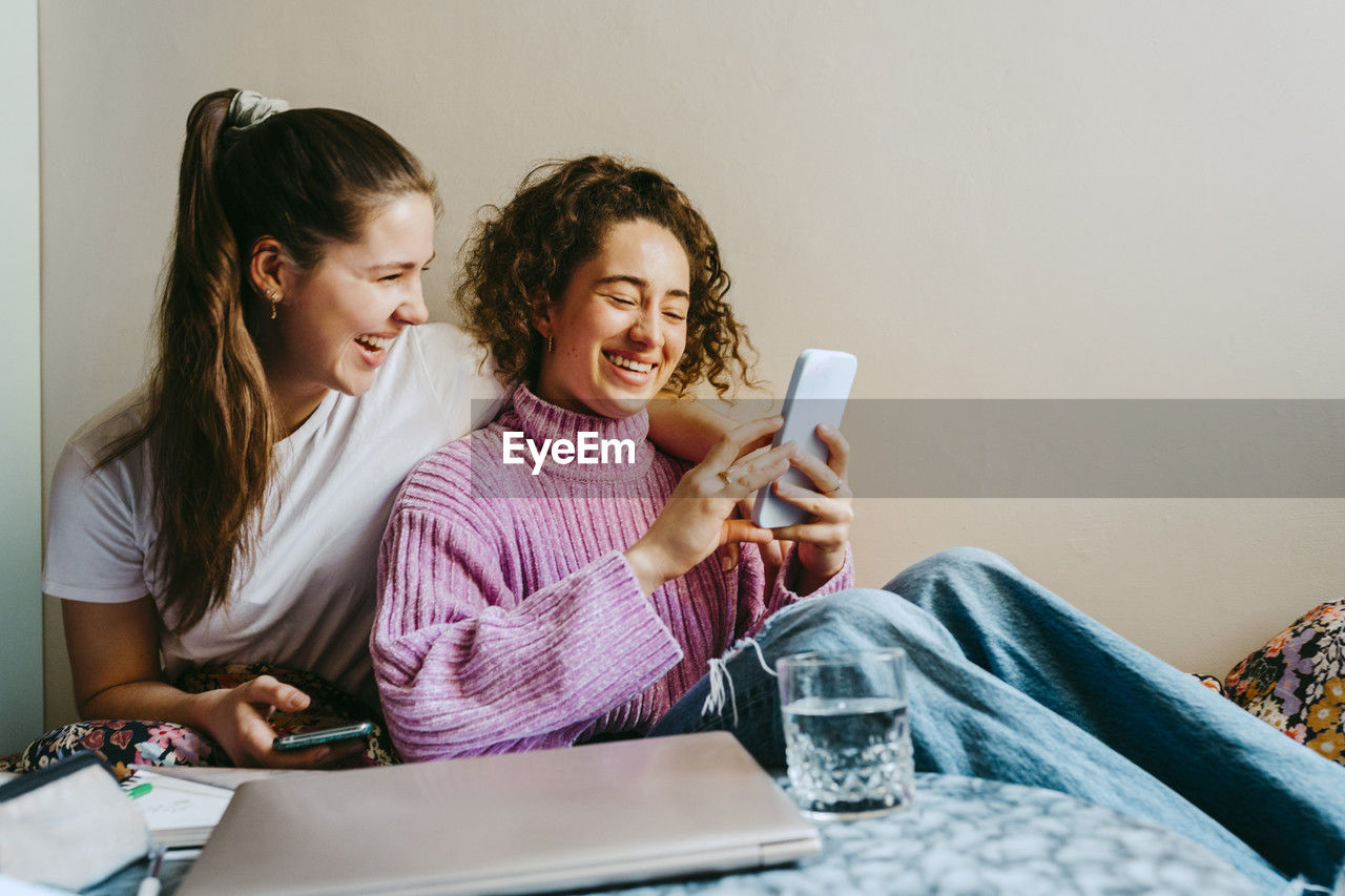 Cheerful young female friends sharing smart phone while sitting at home