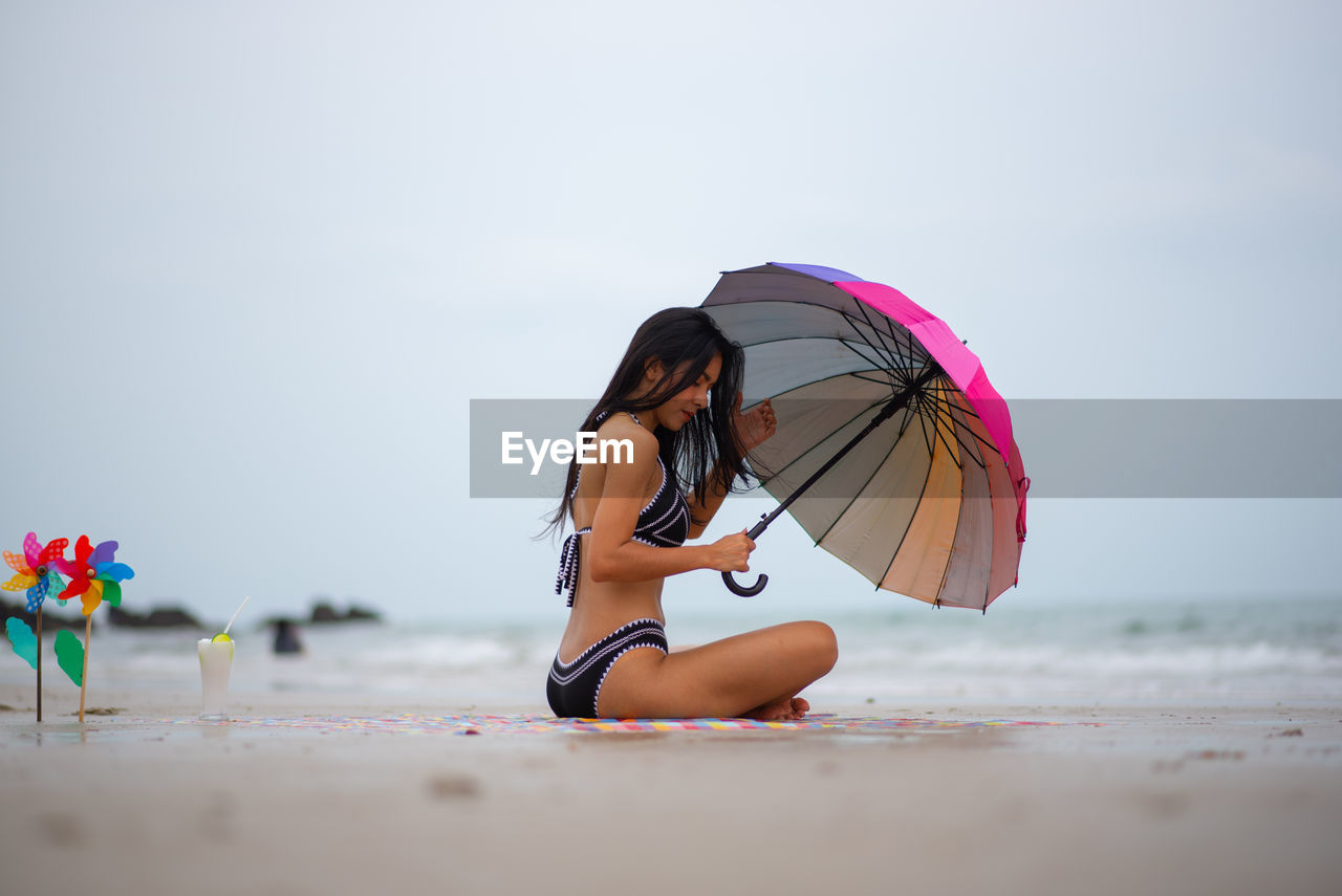 Side view of woman holding umbrella while sitting at beach against sky