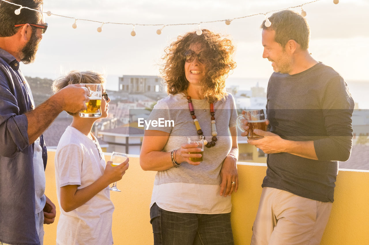 Group of people holding drinks while standing on building terrace during sunset