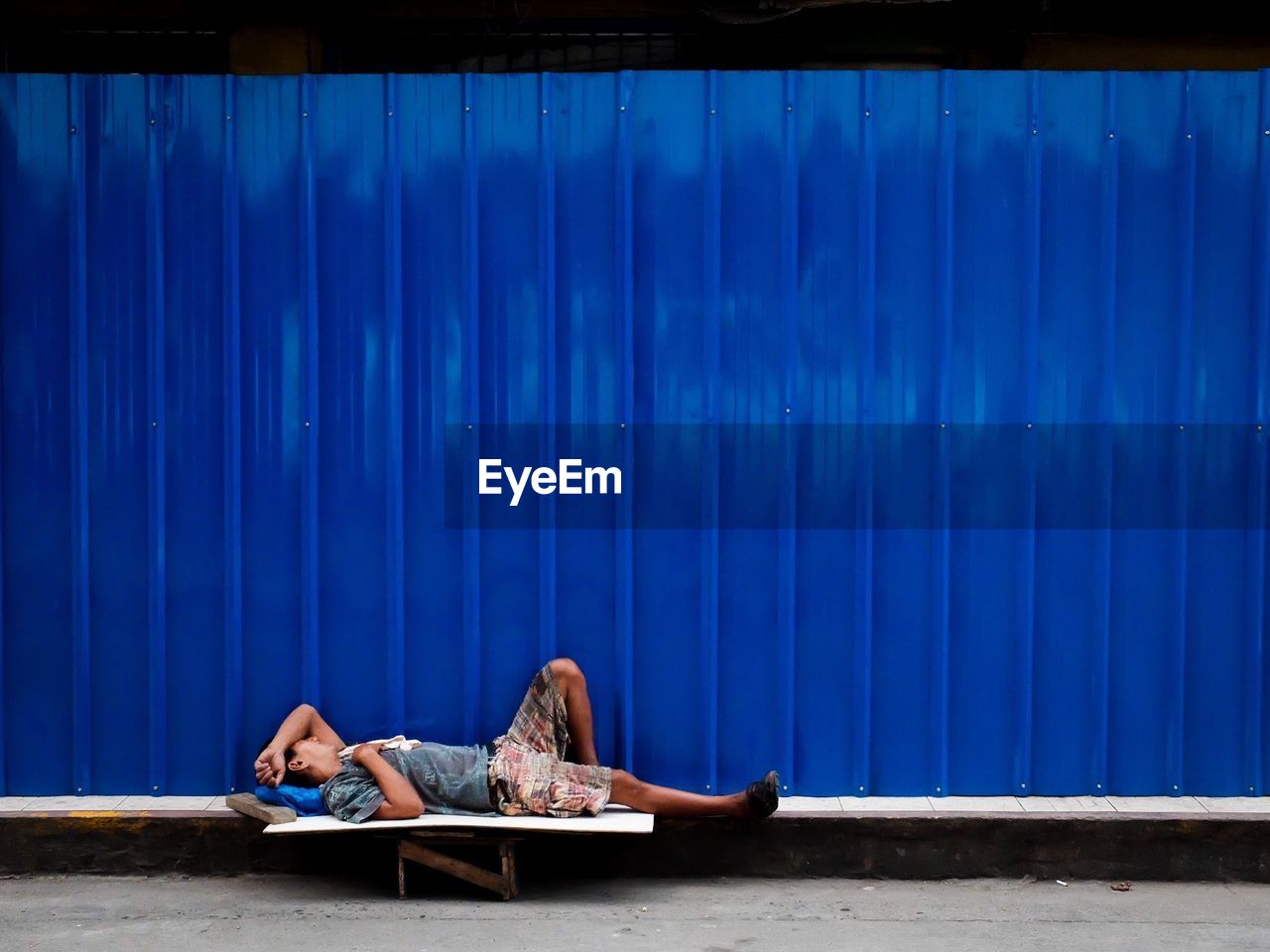 Side view of beggar sleeping against blue metallic wall