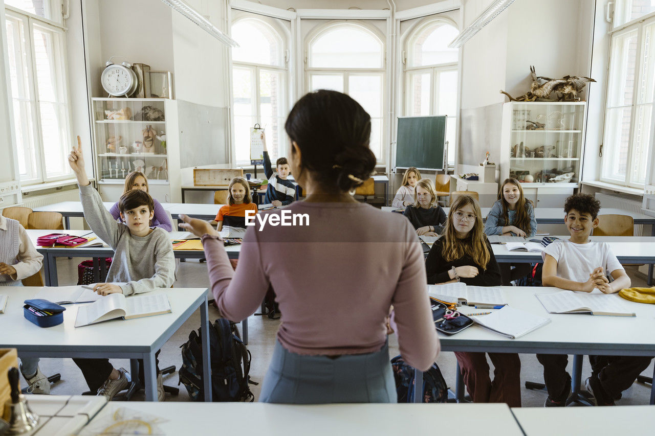 Rear view of female teacher asking students while teaching in classroom