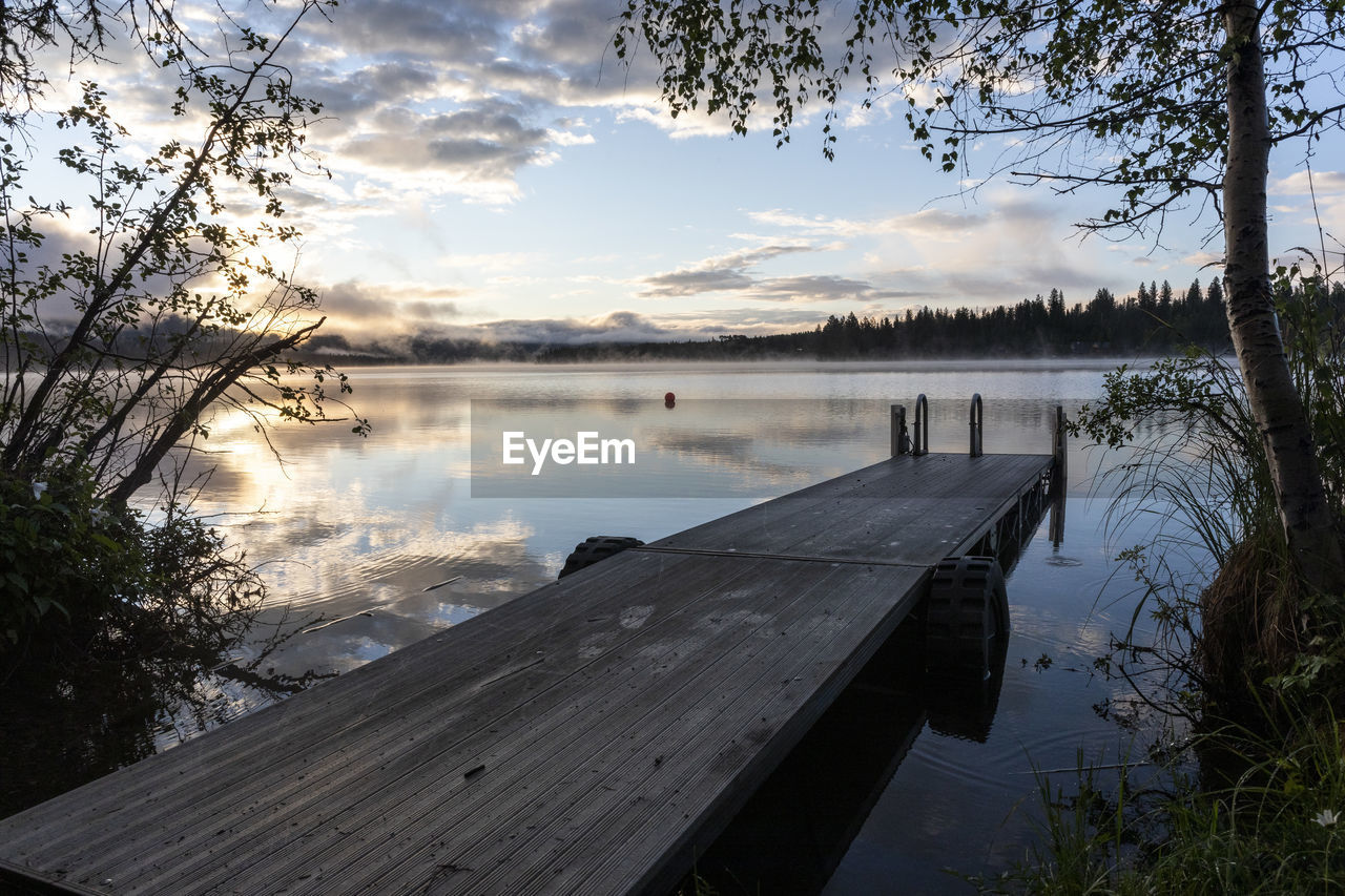 Dock by calm lake and tree during sunrise with reflection of clouds