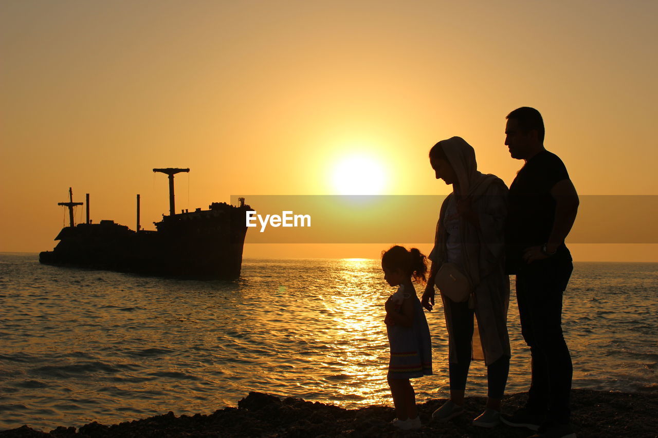 Silhouette family on shore against sky during sunset