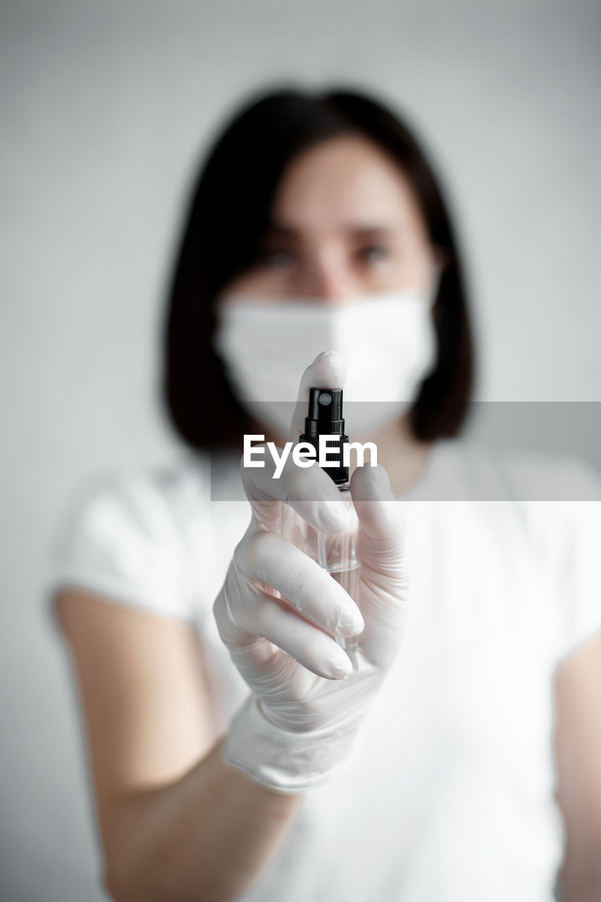 Woman wearing gloves and mask using sanitizer against wall