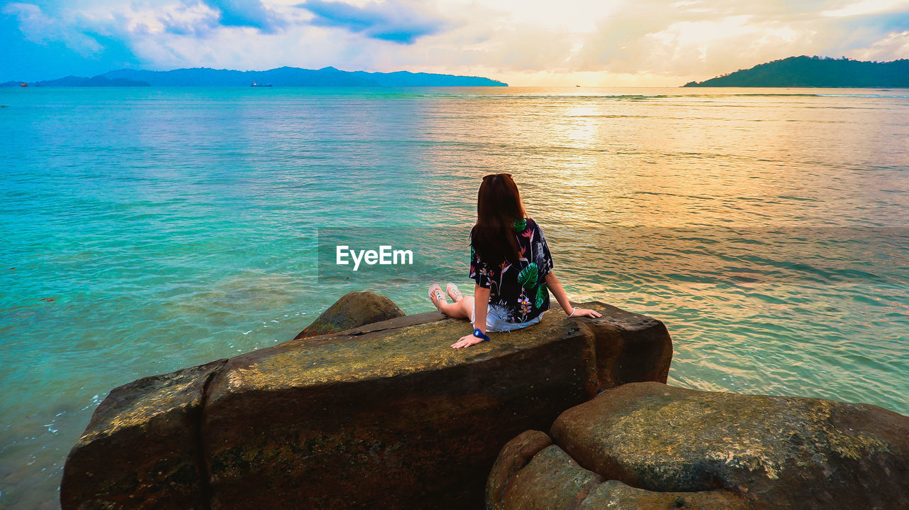 MAN SITTING ON ROCK LOOKING AT SEA SHORE AGAINST SKY