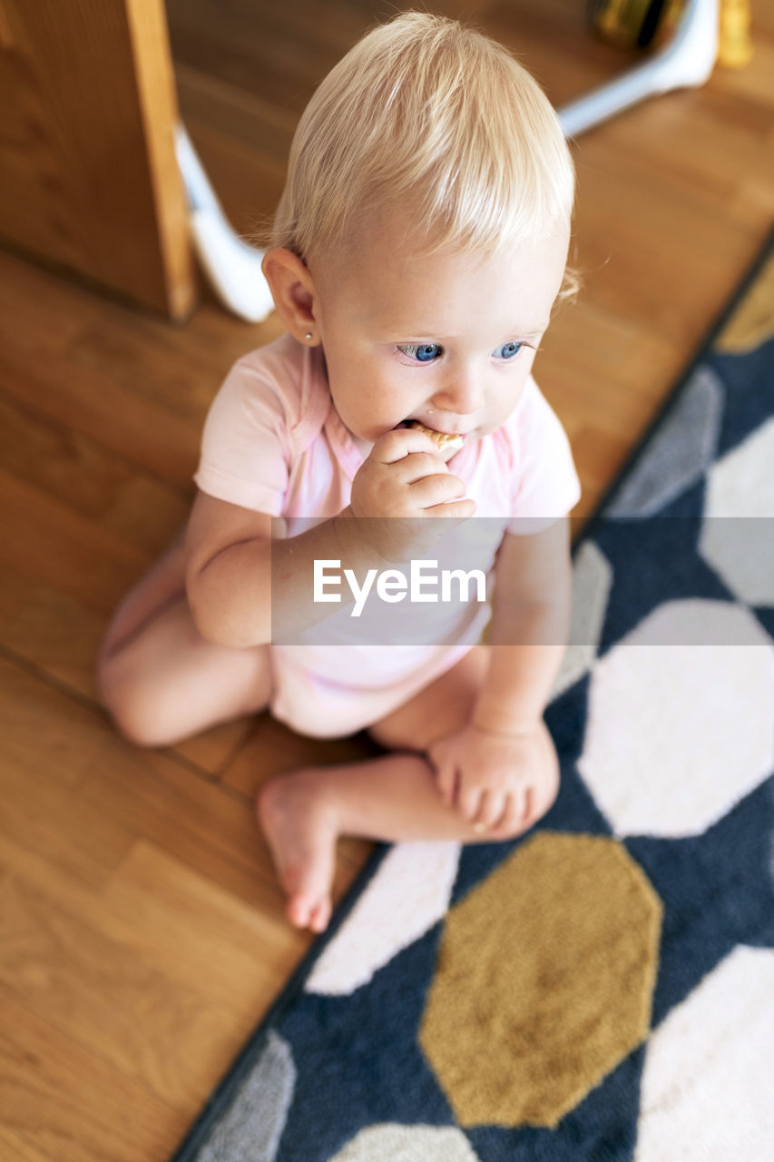 From above of adorable baby girl with blue eyes and blond hair eating cookie while sitting on floor at home