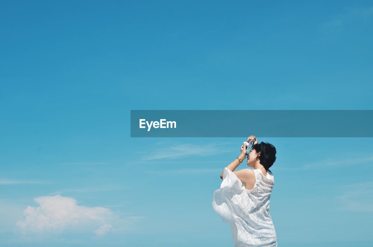 Low angle view of woman photographing against blue sky