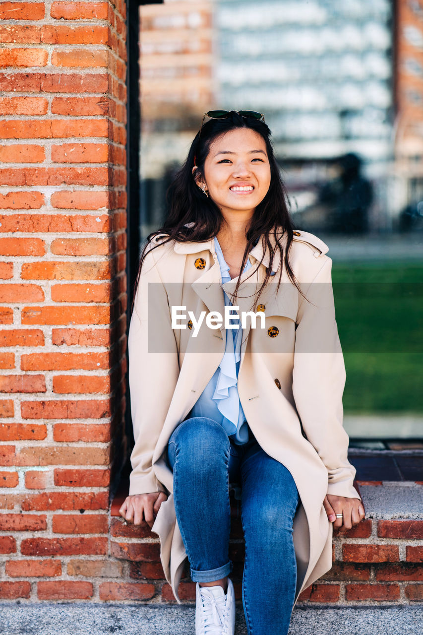 Confident young ethnic lady with long dark hair in stylish coat smiling while sitting on brick wall of aged building and looking at camera