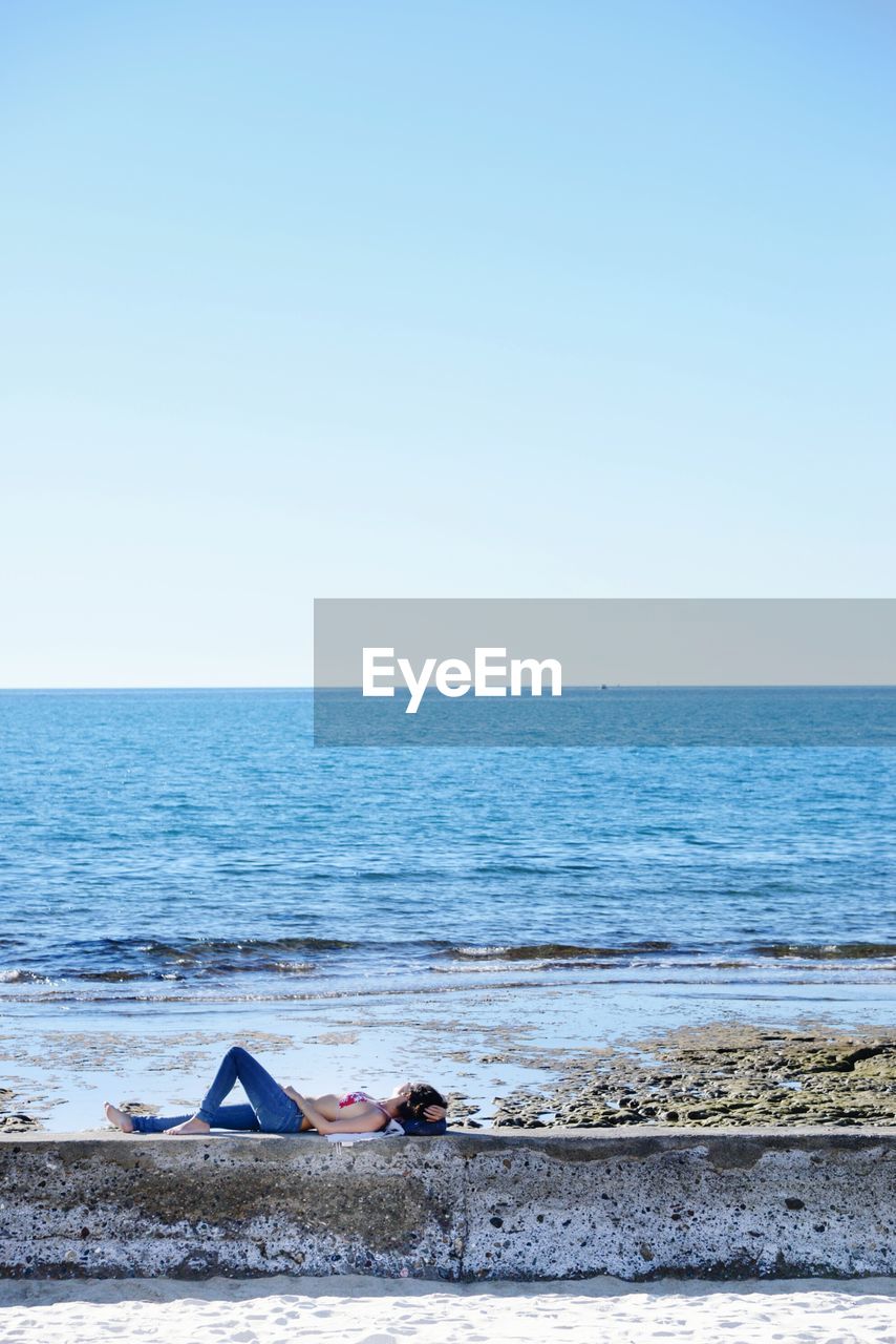 Side view of woman lying on retaining wall at beach against clear blue sky