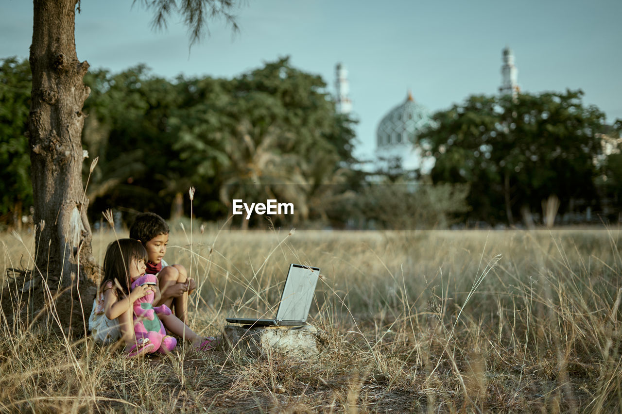 Siblings with laptop sitting on grassy land