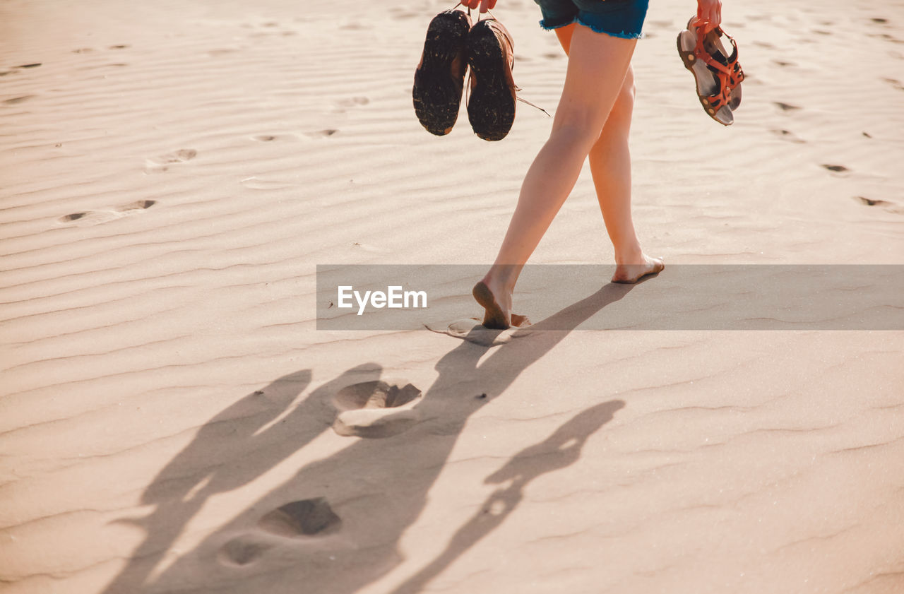 Low section of woman holding shoes while walking on sand at beach