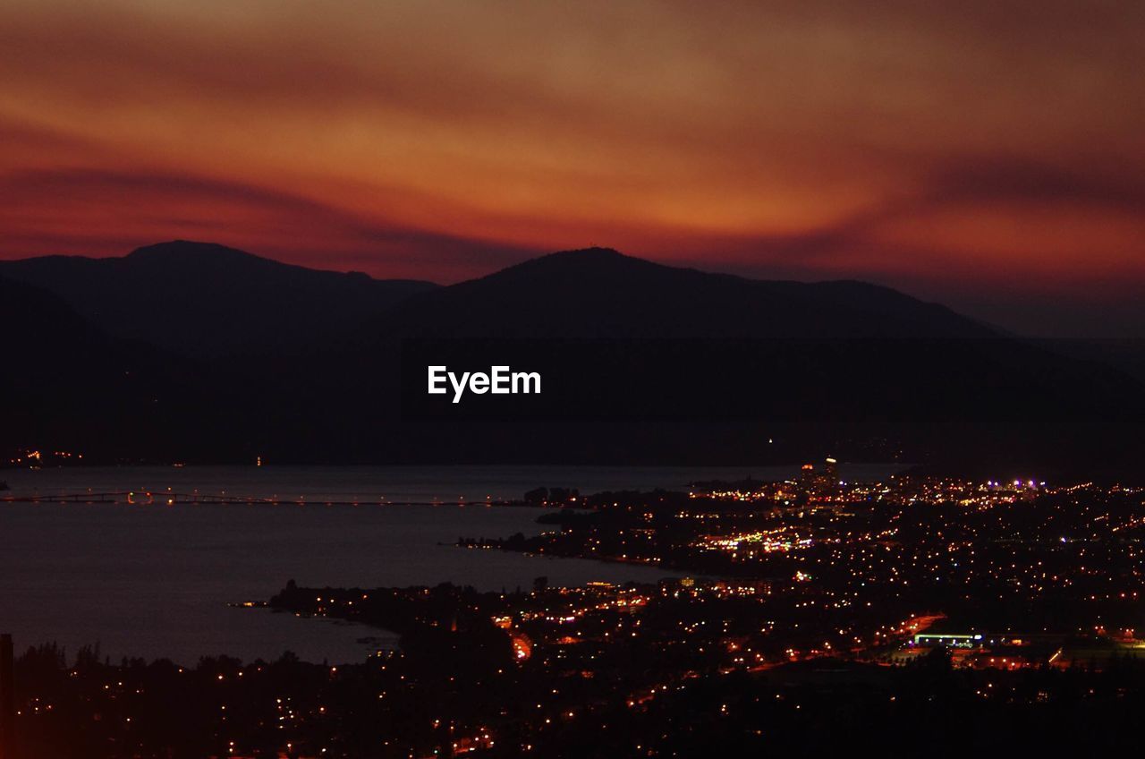 Aerial view of illuminated city by river against silhouette mountains and sky