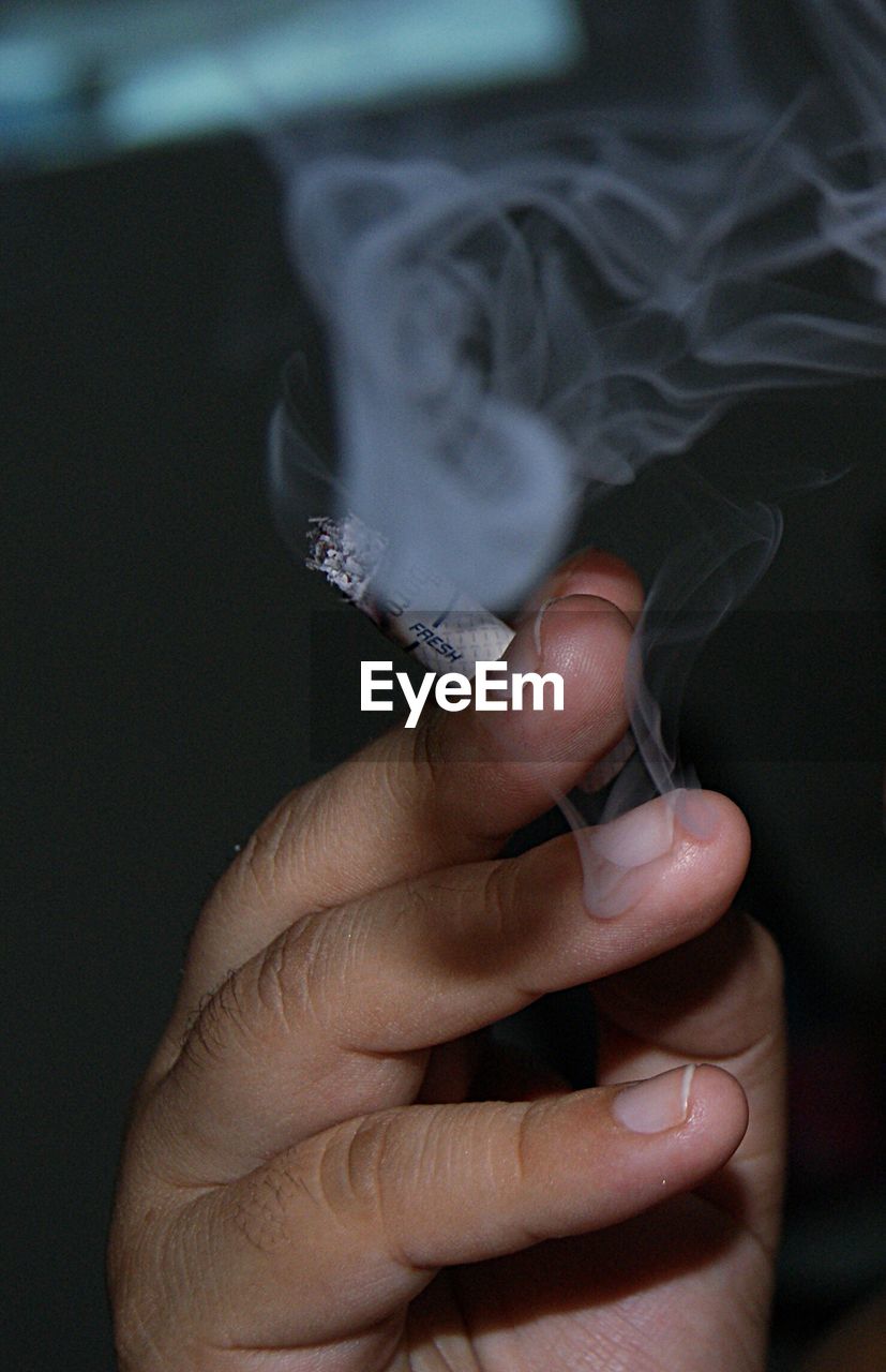 Close-up of hand holding cigarette with smoke in darkroom