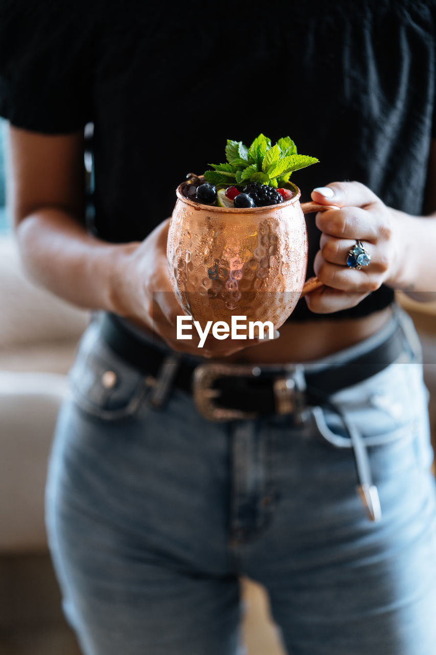 Unrecognizable female carrying metal mug of natural fruit drink with berries and mint leaves on summer day in cafe