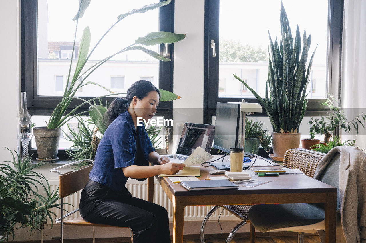 Side view of mature female architect examining documents while sitting at desk in home office