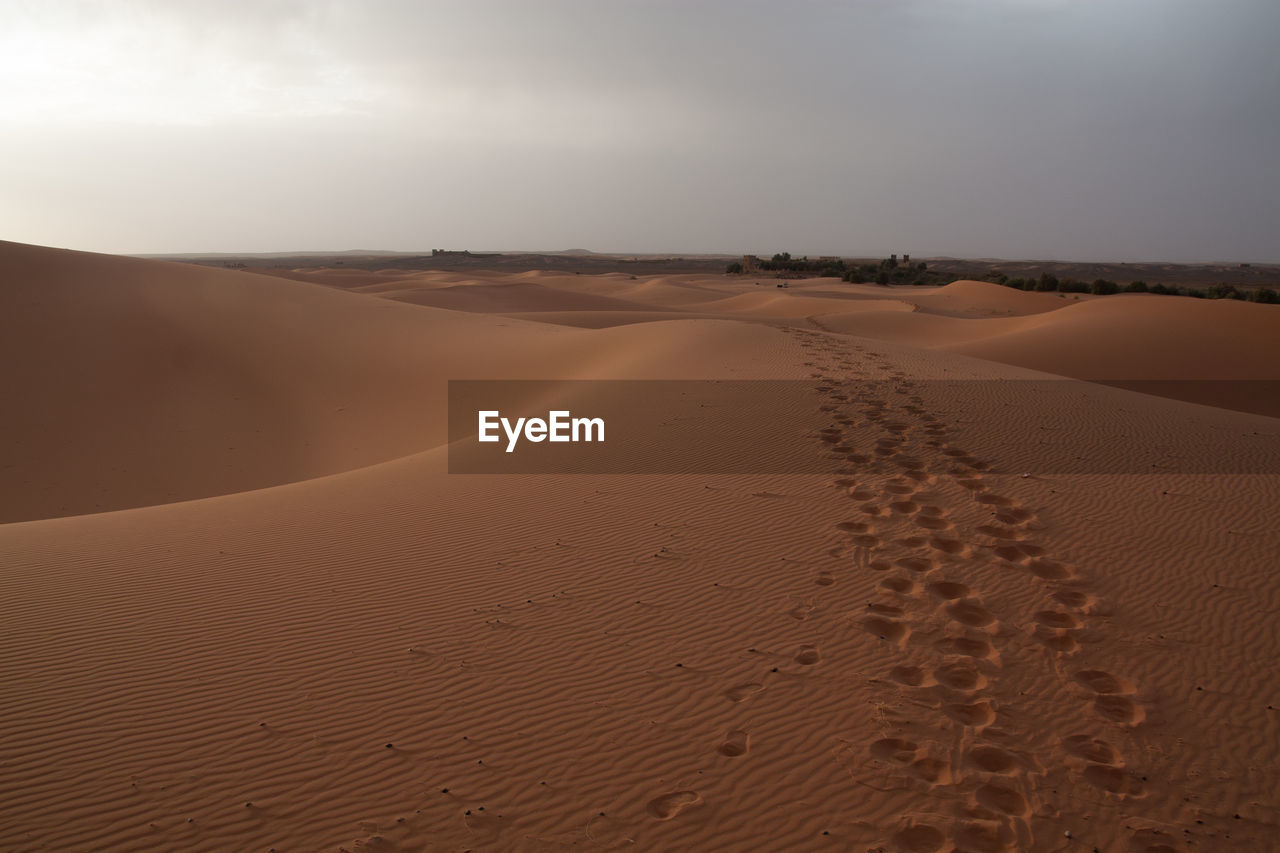 Footprints on sand dune in desert against cloudy sky
