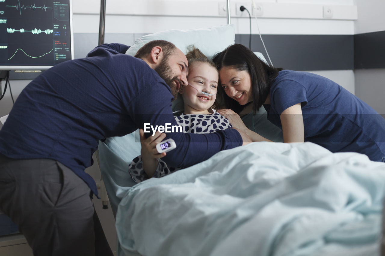 Smiling parents embracing daughter with oxygen tube lying on bed at hospital ward