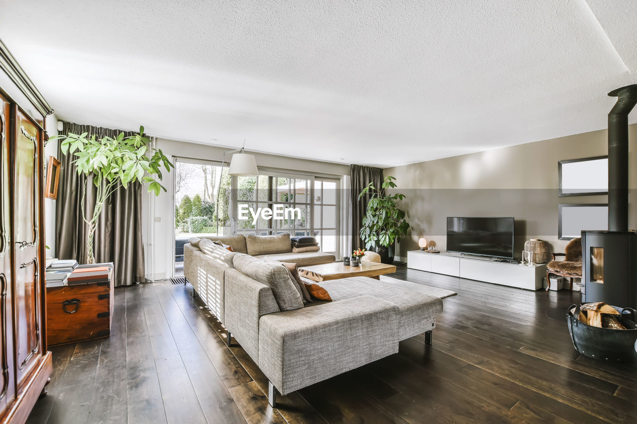 Comfortable sofa placed on rug with table against wall with tv in stylish living room with green plants at window