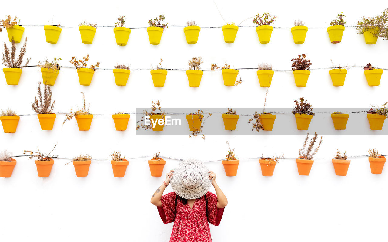 Anonymous female tourist covering face with straw hat near white wall with different potted plants