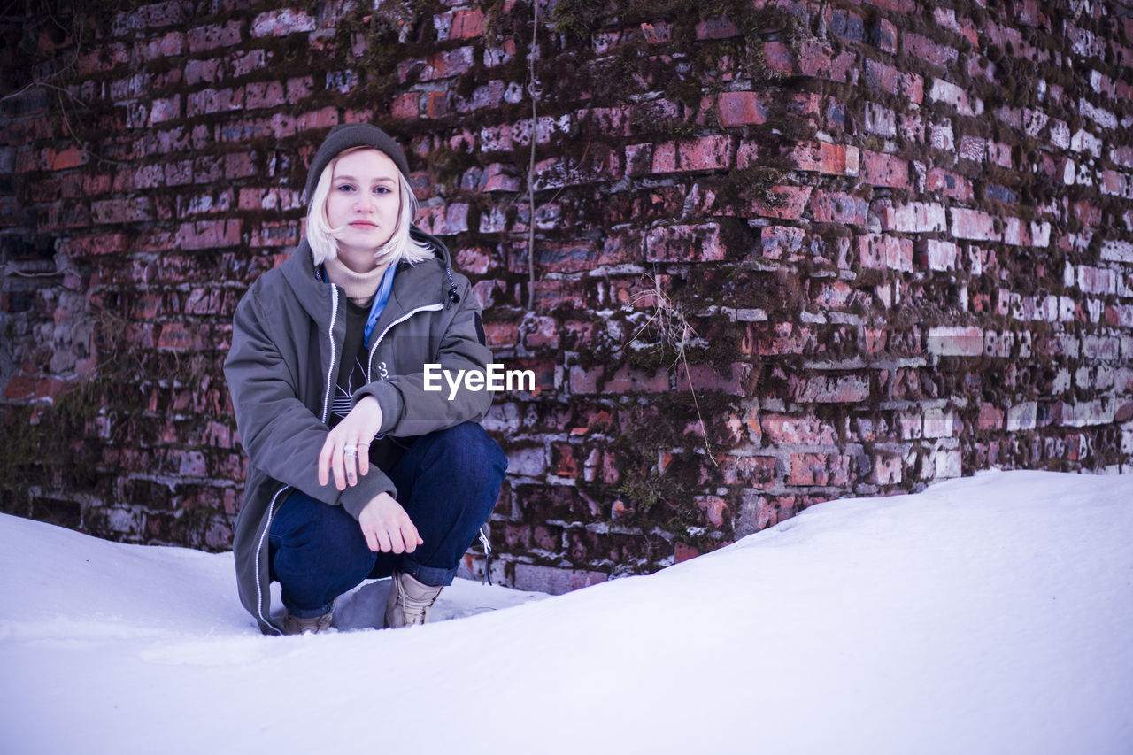 Portrait of woman crouching against brick wall during winter