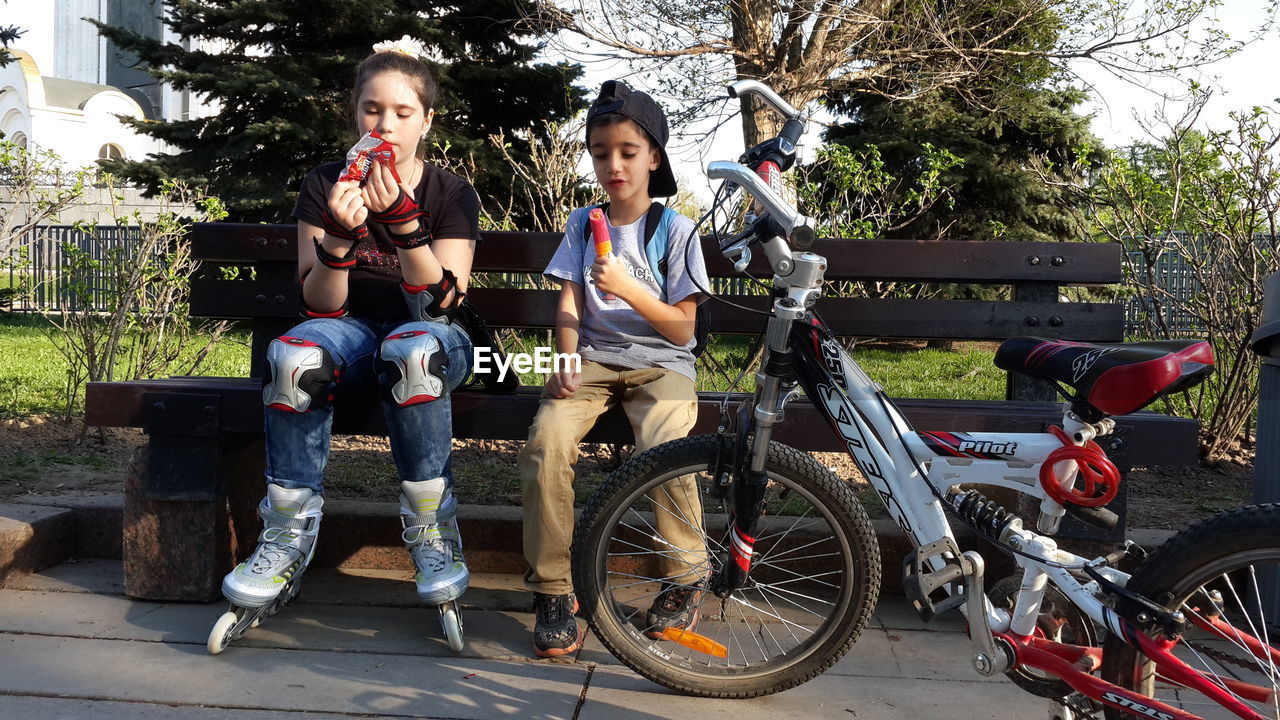 Full length of siblings having food while sitting on bench at park