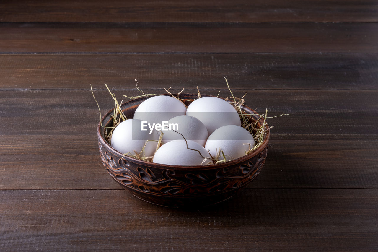 White raw chicken eggs lie on hay in a burnt clay bowl on the surface of their natural dark wood. 