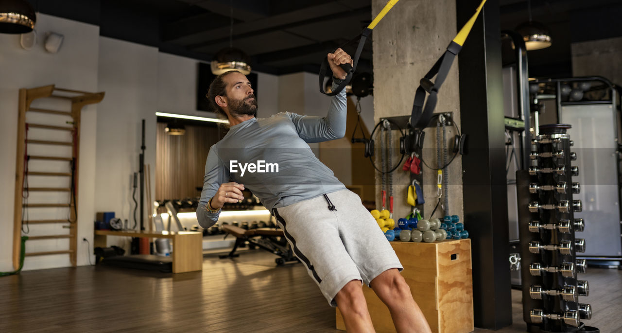 Side view of concentrated muscular male athlete practicing suspension training with trx ropes in modern gym