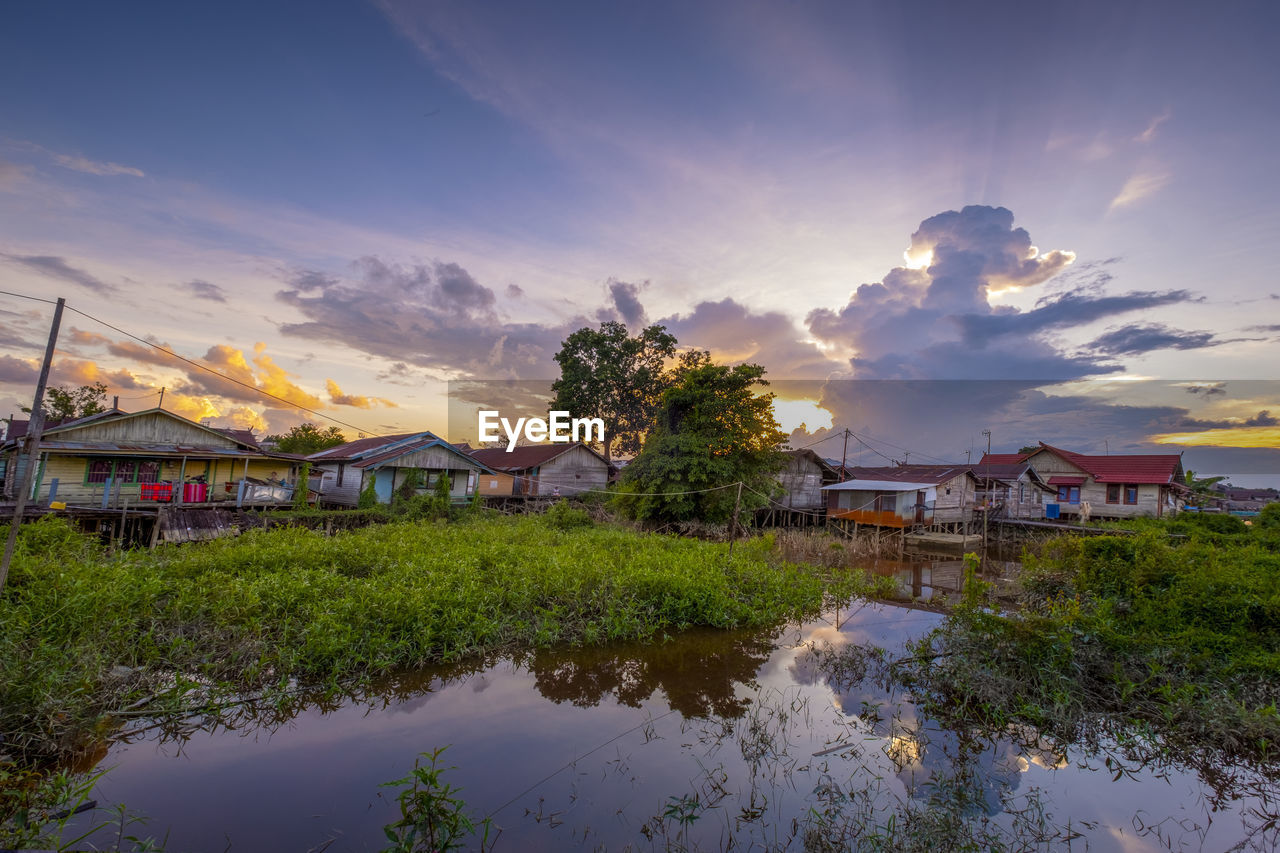 Houses by river and buildings against sky at sunset