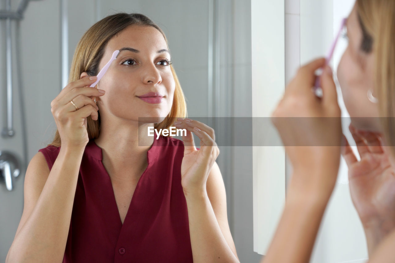 Close up of latin woman shaving her eyebrows with razor in the mirror at home