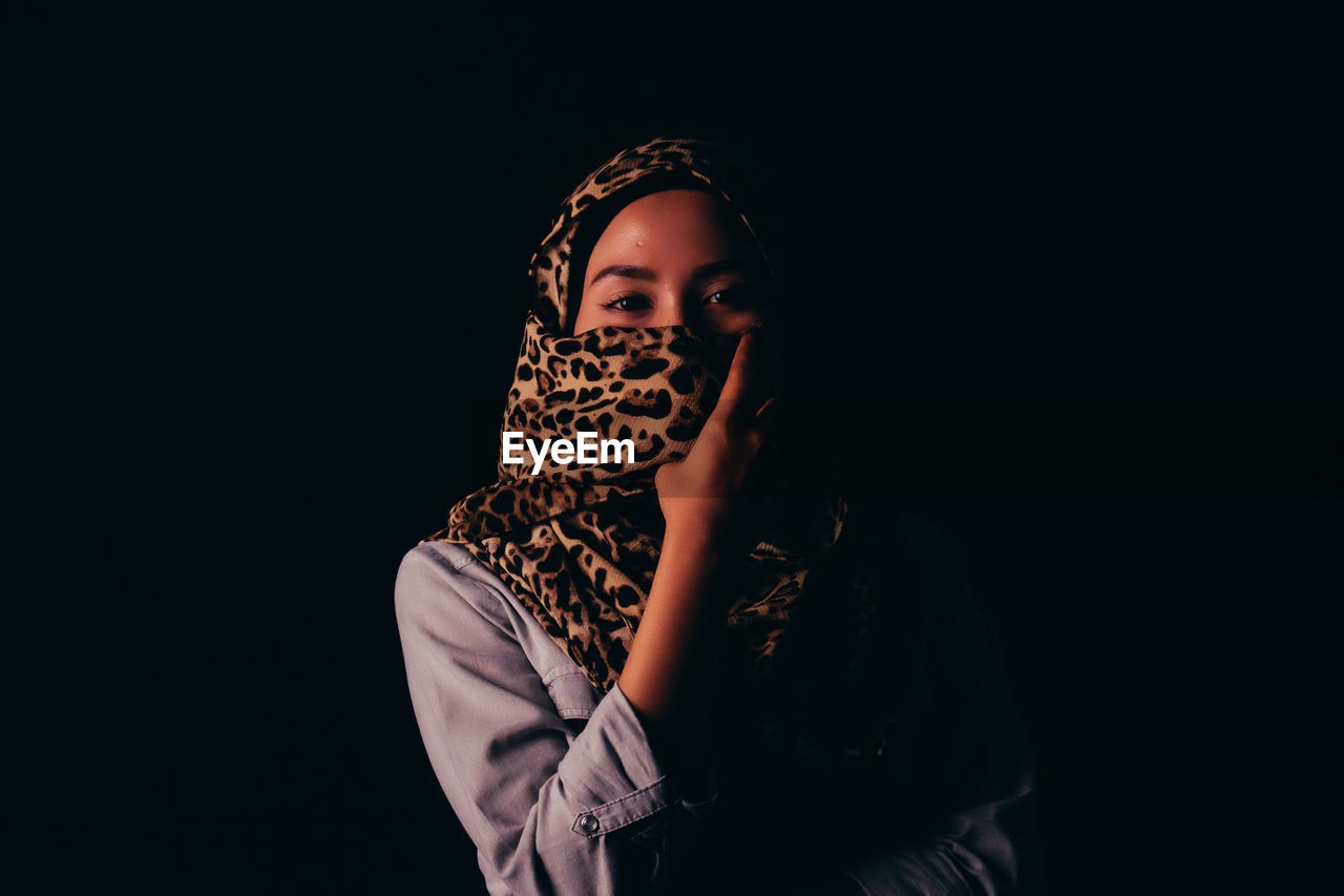 Portrait of young woman covering face with headscarf against black background