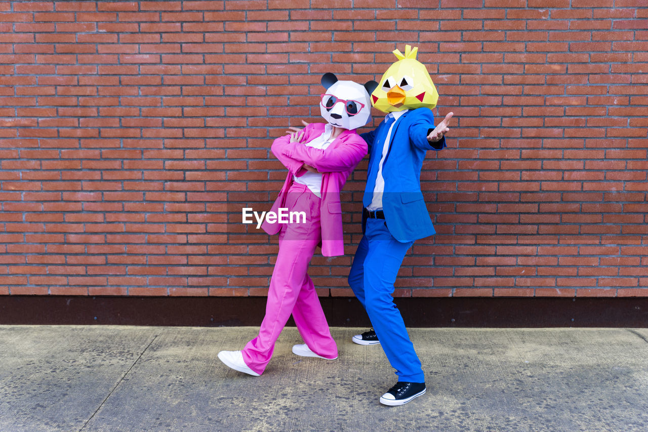 Man and woman wearing vibrant suits and animal masks posing together in front of brick wall