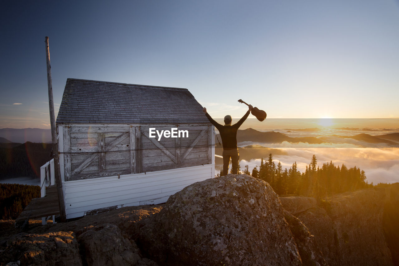 Rear view of man holding guitar while standing by log cabin on mountain during sunset