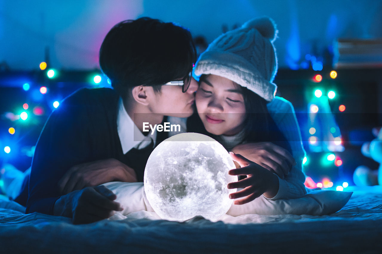 Boyfriend kissing girlfriend by illuminated moon light decoration on bed