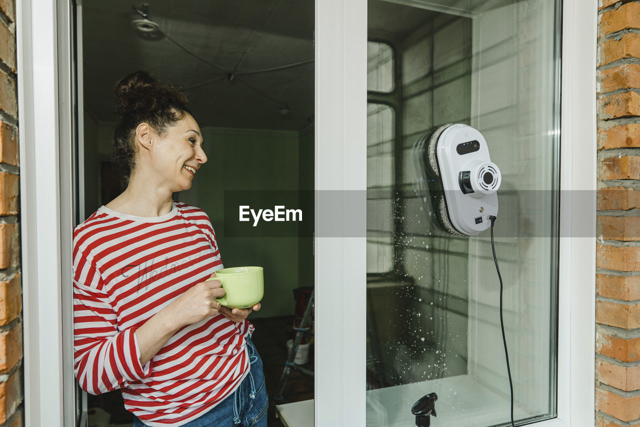 Happy woman with coffee cup cleaning window through robot washer