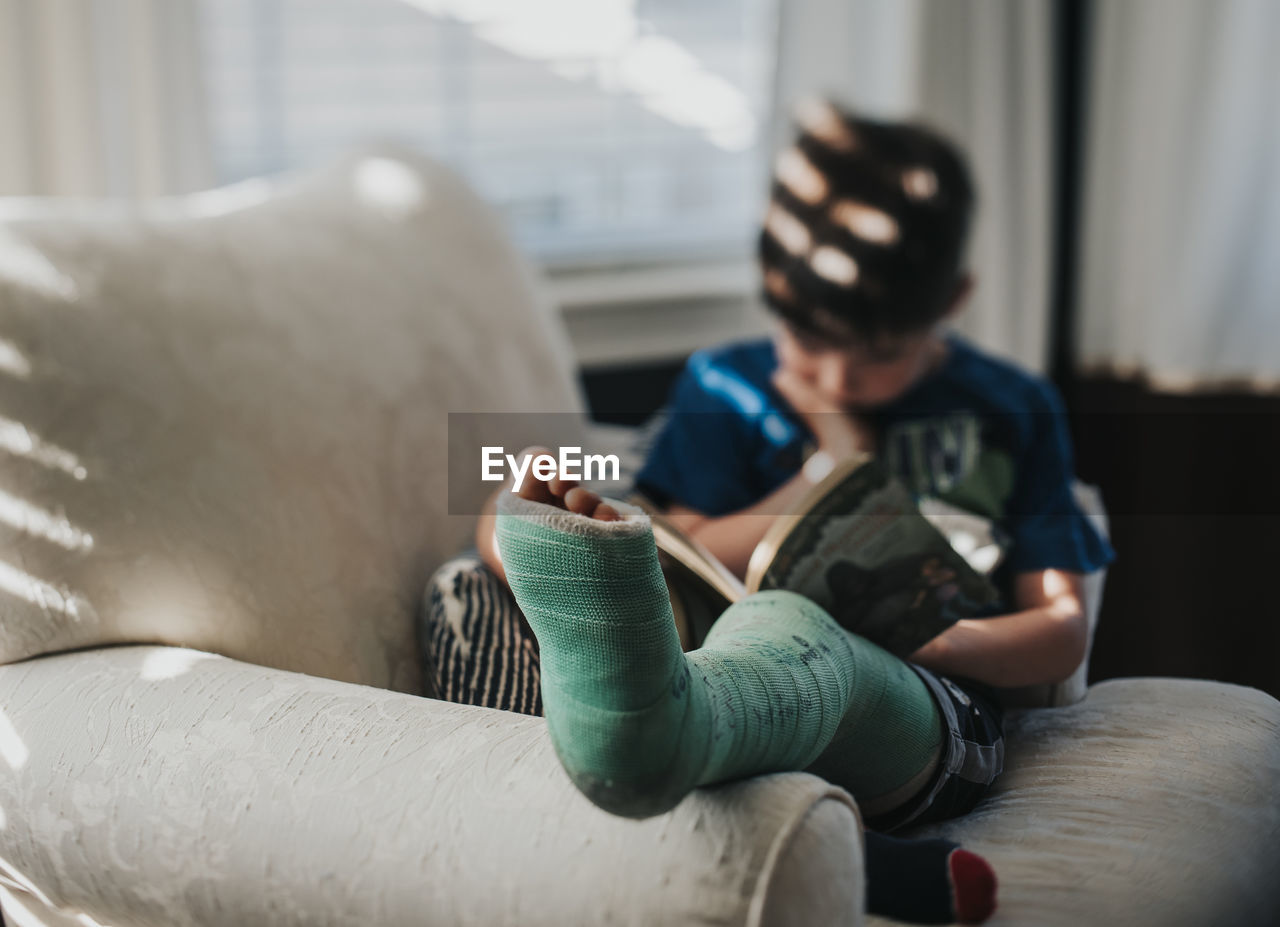 Boy with broken leg reading book while relaxing on armchair at home