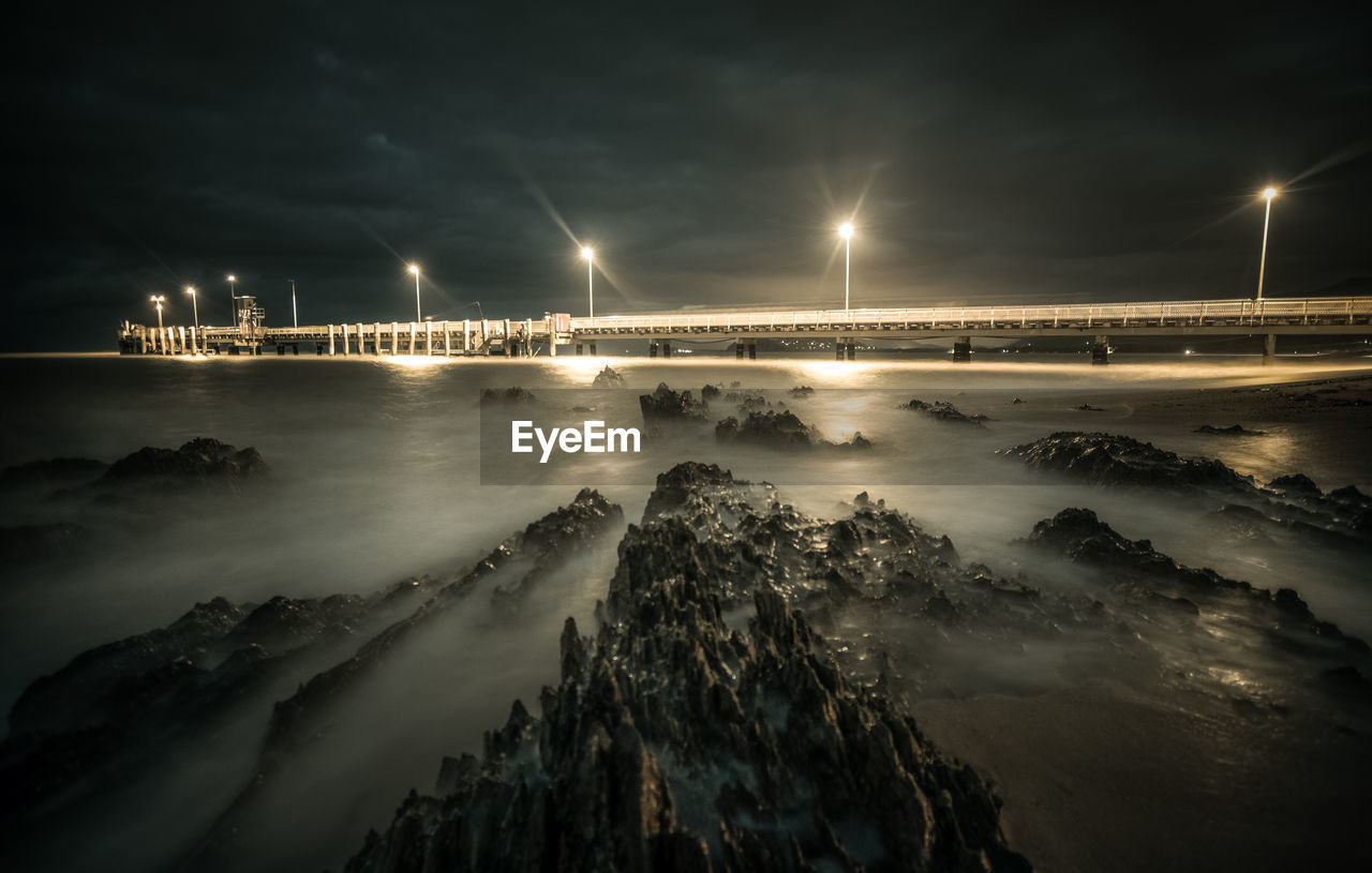 High angle view of rocks against illuminated pier over sea at night