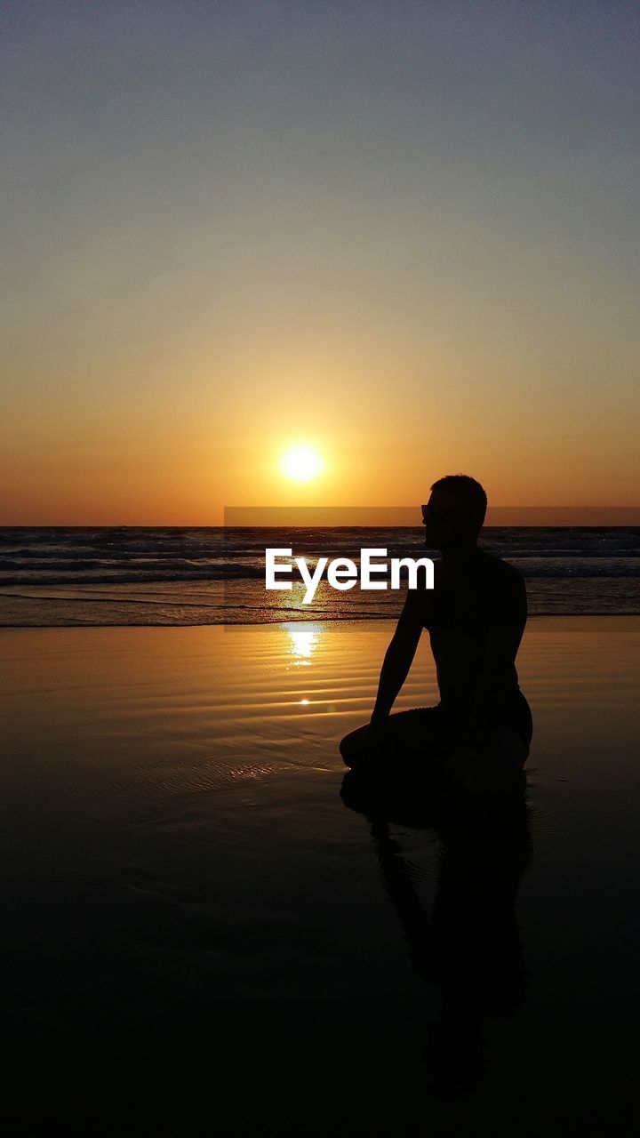 SILHOUETTE MAN SITTING ON BEACH AGAINST SKY AT SUNSET