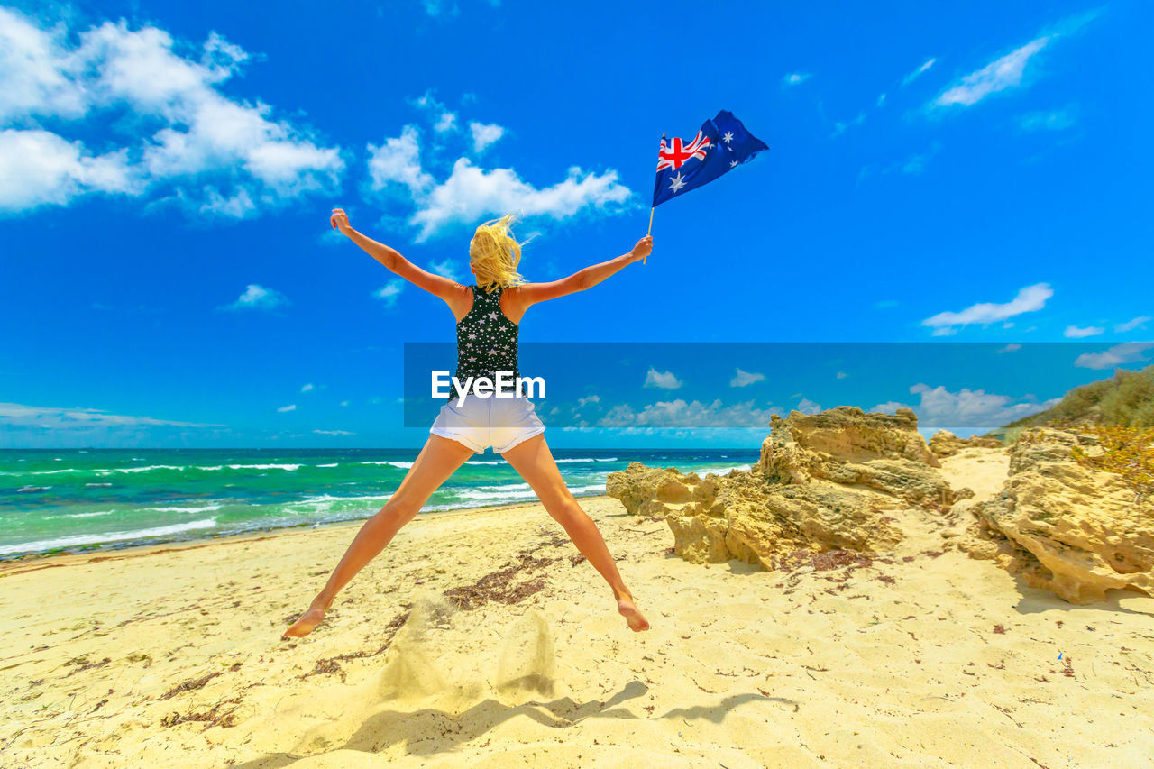 Rear view full length of woman holding australian flag jumping at beach